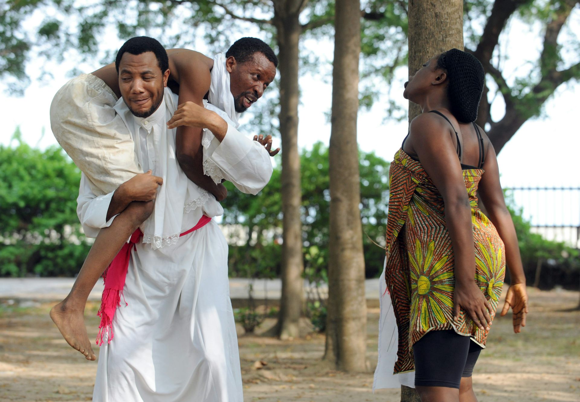 three actors on an outdoor stage, trees in the background. A man carries a yelling man on his shoulders, moving away from a woman yelling back at the man.
