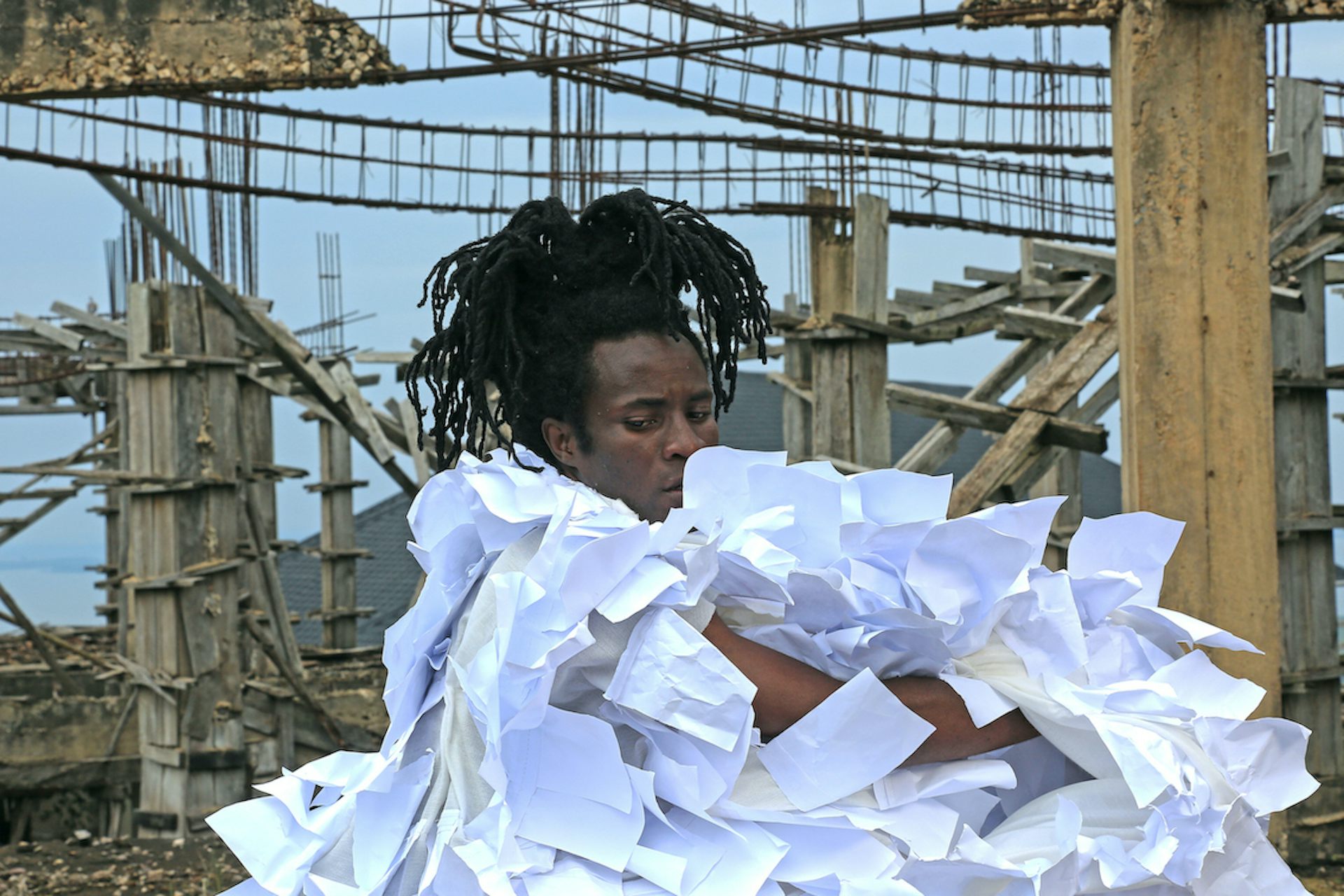 A man at a construction site, his hair a cascade of dreadlocks, he is wrapped in a cloak made of multiple sheets of white paper.