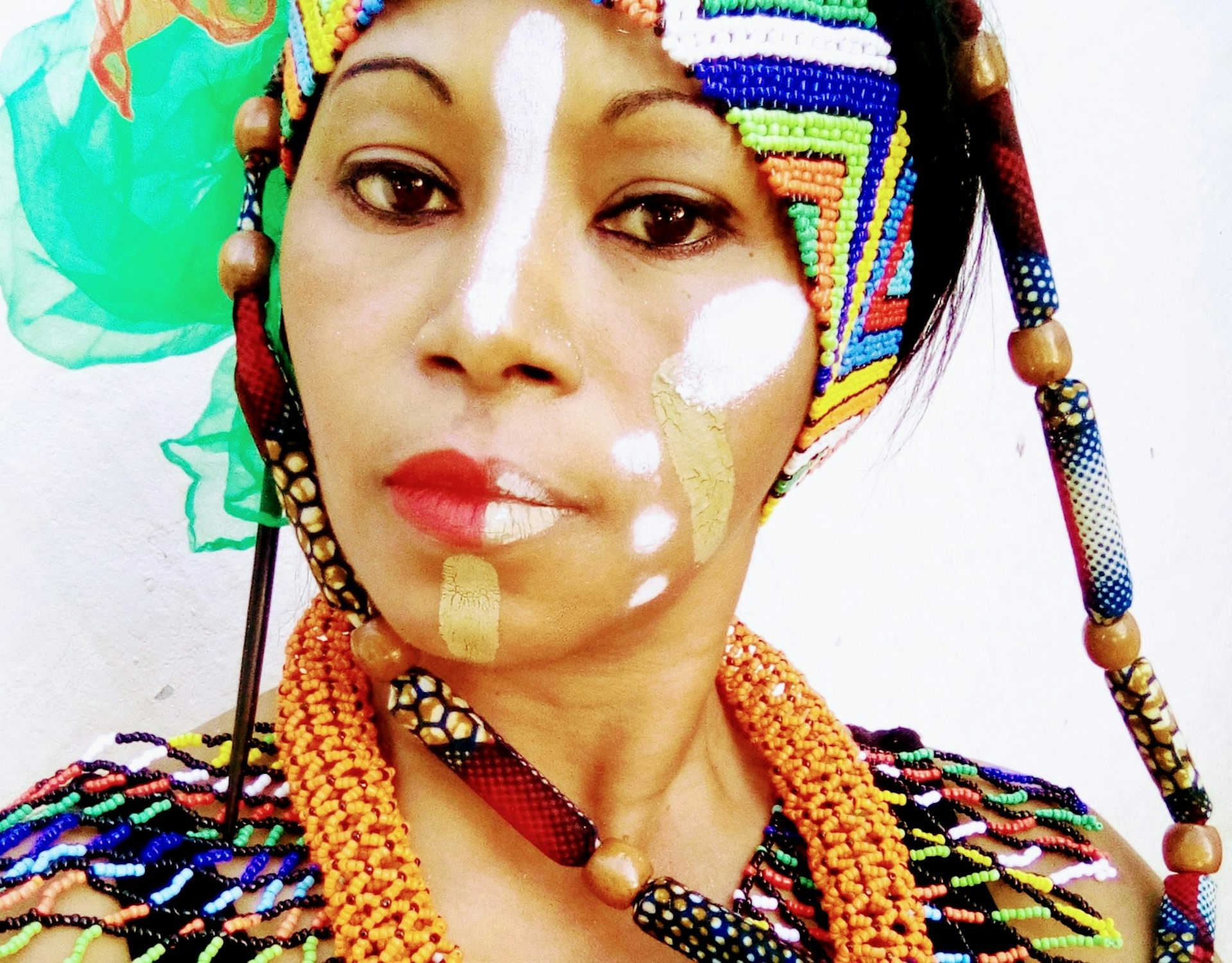 a woman's face with ethnic make-up smears in tan and white, a brightly coloured scarf in her hair matches her jewellery.